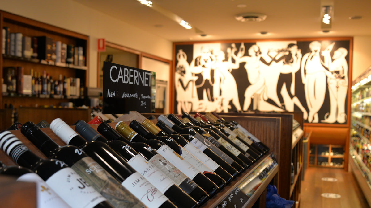 A wine display shelf inside a wine store, showing multiple bottles of red wine with labels including Cabernet, set against a warm interior with shelving and a large black-and-white wall mural.