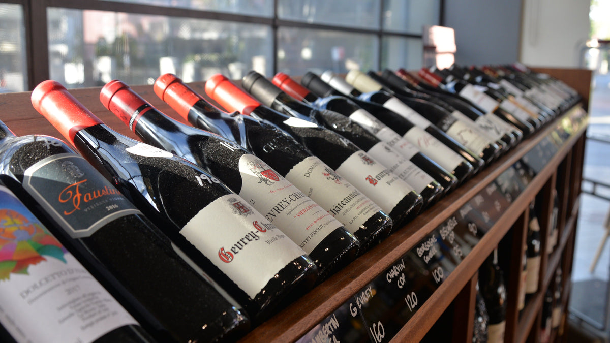 A wooden wine rack filled with rows of wine bottles, photographed in a real indoor setting with large windows in the background. Several bottle labels are visible, including a prominent Faulkner label and a G.